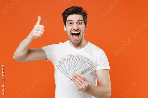 Obraz na plátně Excited laughing screaming young man in basic casual empty blank white t-shirt hold fan of cash money in dollar banknotes showing thumb up isolated on bright orange colour background studio portrait