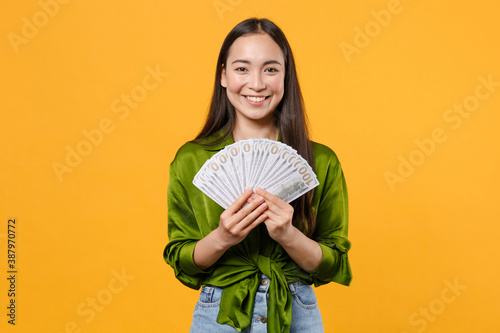 Fotografie Smiling young brunette asian woman wearing basic green shirt standing hold fan of cash money in dollar banknotes looking camera isolated on bright yellow colour background, studio portrait
