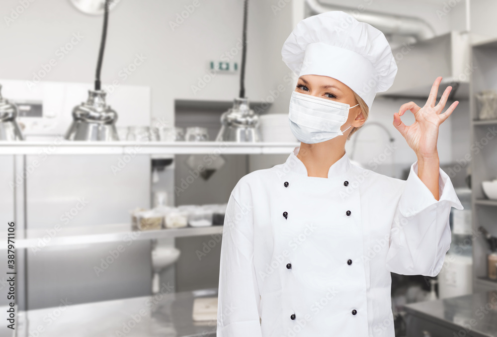 cooking, culinary and health concept - female chef in toque wearing face protective medical mask for protection from virus disease showing ok hand sign over restaurant kitchen background