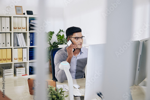 Indian entrepreneur in glasses sitting at office desk and talking on phone with colleague