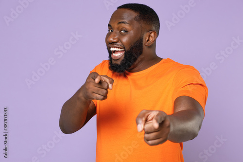 Cheerful excited young african american man 20s wearing basic casual orange blank empty t-shirt standing pointing index fingers on camera isolated on pastel violet colour background, studio portrait.