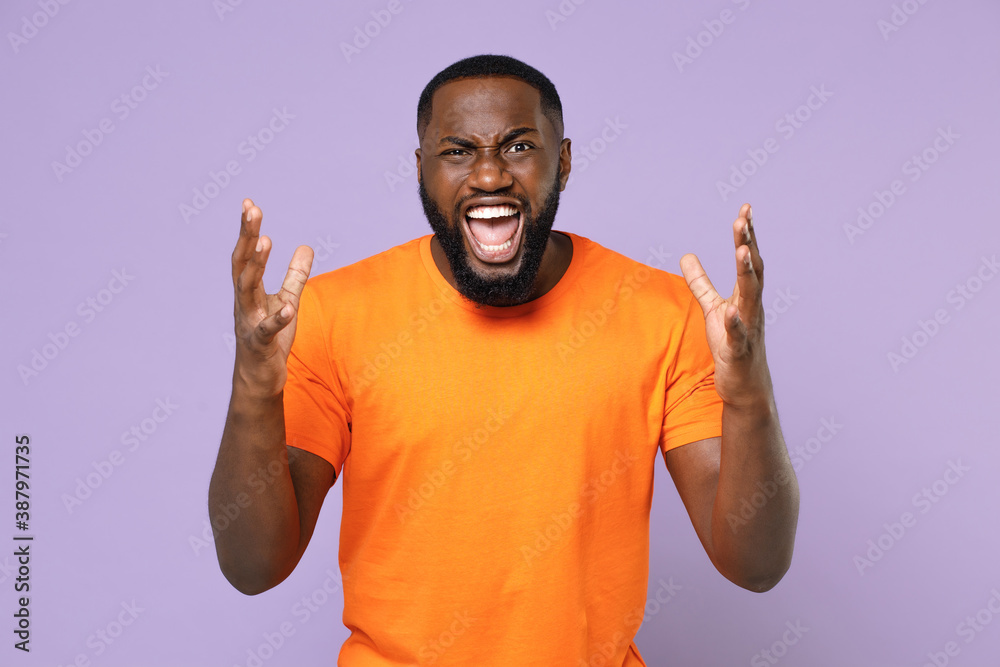 Fototapeta premium Angry irritated young african american man 20s wearing basic casual blank empty orange t-shirt standing spreading hands screaming swearing isolated on pastel violet colour background studio portrait.