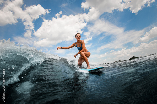 Athletic and active woman riding on wave sitting on surfboard against blue sky
