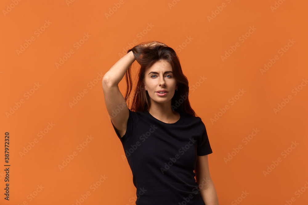 Flirting. Confident charming playful coquettish caucasian woman in black t-shirt raises hair, smiling shyly at camera. Isolated over an orange background. Flirting. Confident charming playful coquettish caucasian woman in black t-shirt raises hair, smiling shyly at camera. Isolated over an orange background.