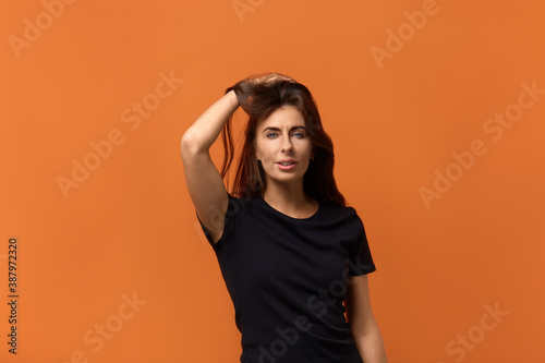 Flirting. Confident charming playful coquettish caucasian woman in black t-shirt raises hair, smiling shyly at camera. Isolated over an orange background. © 5M