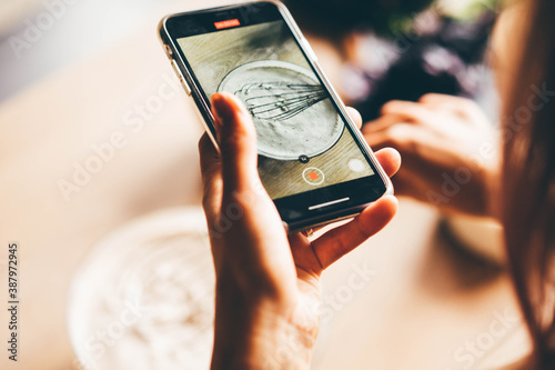 Skilled culinary blogger holds smartphone and makes video of mixing dish products in bowl with metal whisk on brown wooden table at home back upper view photo