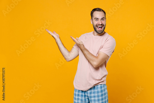 Funny excited young bearded man in pajamas home wear point hands aside up on mock up copy space while resting at home isolated on yellow background studio portrait. Relax good mood lifestyle concept.