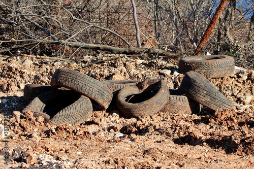 Bunch of old heavily used truck tyres covered with dirt left at edge of local construction site for recycling surrounded with rocks and tree branches on warm sunny winter day