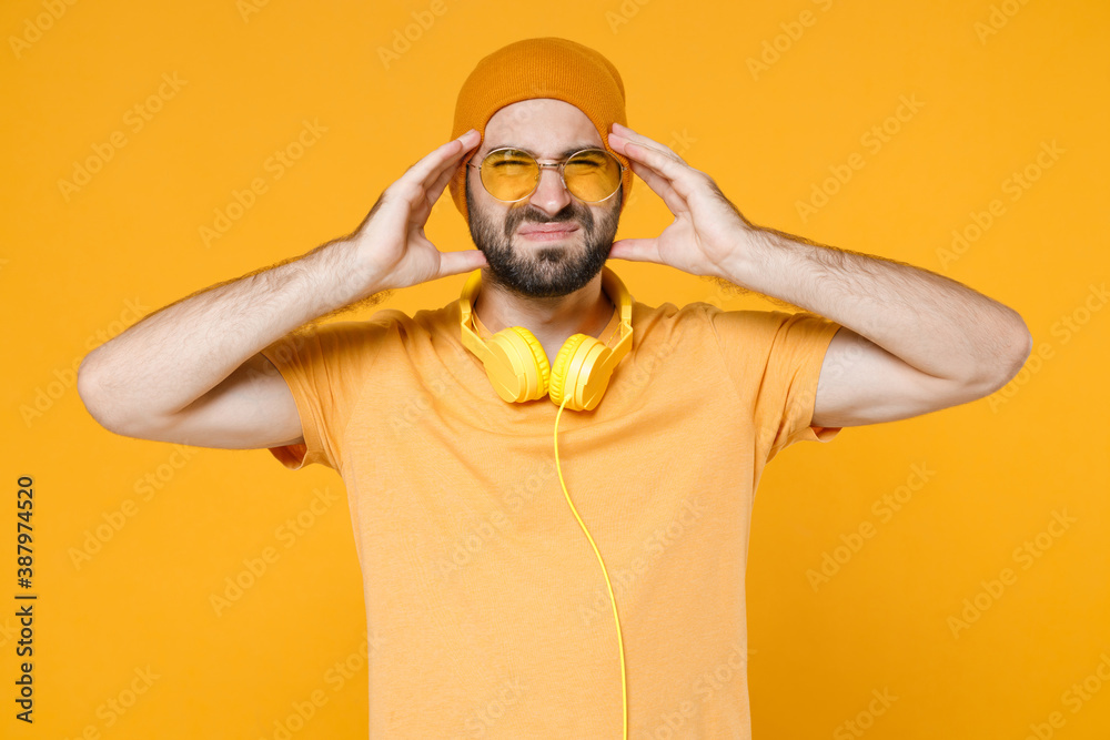 Dissatisfied sick tired young bearded man 20s wearing basic casual t-shirt headphones eyeglasses hat standing put hands on head having headache isolated on bright yellow background studio portrait. Dissatisfied sick tired young bearded man 20s wearing basic casual t-shirt headphones eyeglasses hat standing put hands on head having headache isolated on bright yellow background studio portrait.