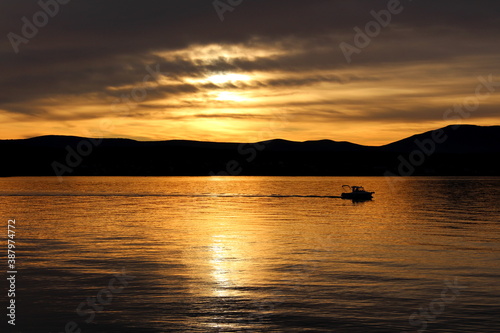 Small fishing boat navigating across the bay and calm sea at beautiful sunset with mountains and cloudy sky in background