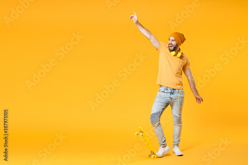 Full length of cheerful laughing young man wearing basic casual t-shirt headphones hat having fun with skateboard rising spreading hands isolated on bright yellow colour background, studio portrait.