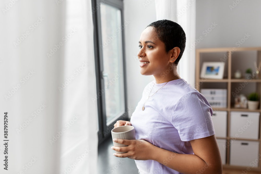 people and leisure concept - happy young african american woman with coffee cup looking out of window at home people and leisure concept - happy young african american woman with coffee cup looking out of window at home