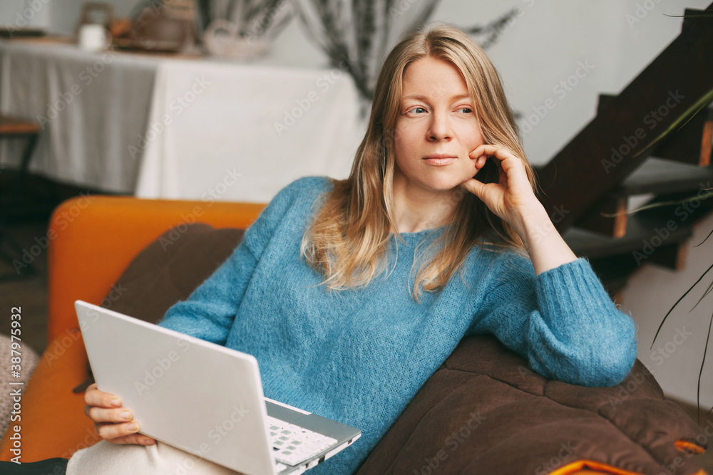 Fototapeta premium Pensive beautiful girl sitting on the couch with a laptop. Young woman working at home using laptop. Work online, freelance, social distance