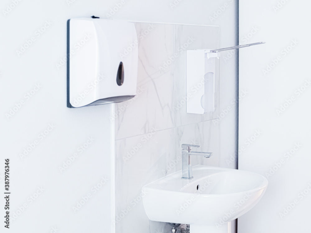 Fototapeta premium Bathroom with soap dispenser, white sink with metal faucet, and plastic tissue paper box, towel hanging on white wall. Light interior