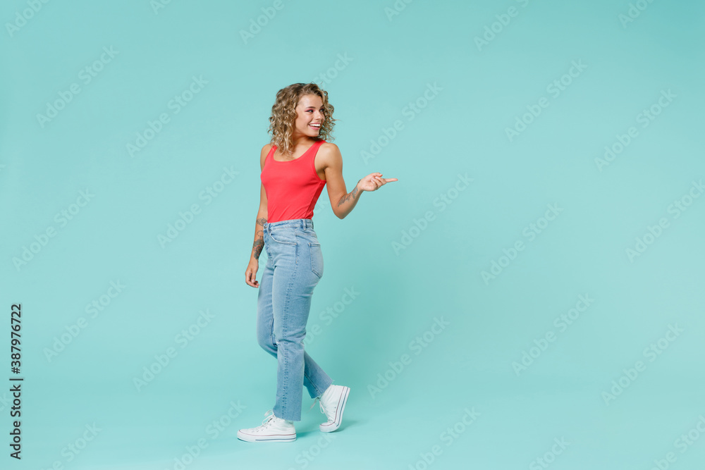 Full length side view of smiling young blonde woman 20s in pink casual tank top jeans standing pointing index finger aside on mock up copy space isolated on blue turquoise background, studio portrait. Full length side view of smiling young blonde woman 20s in pink casual tank top jeans standing pointing index finger aside on mock up copy space isolated on blue turquoise background, studio portrait.