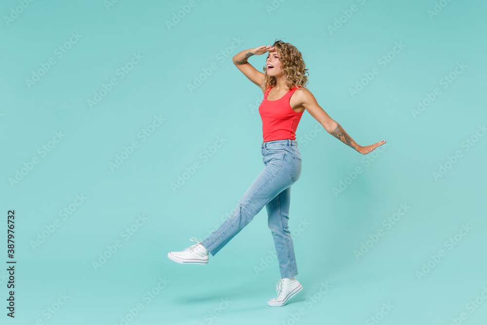 Full length side view of shocked young blonde woman 20s wearing pink tank top jeans standing holding hand at forehead looking far away distance isolated on blue turquoise background, studio portrait.