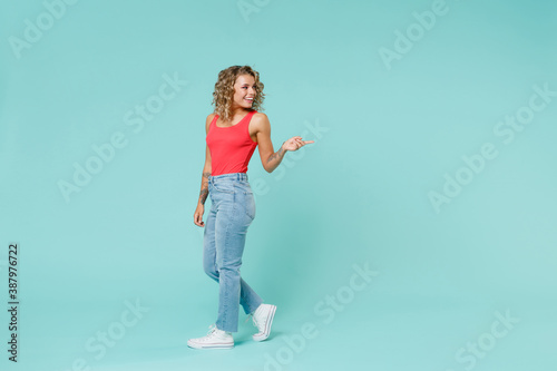 Full length side view of smiling young blonde woman 20s in pink casual tank top jeans standing pointing index finger aside on mock up copy space isolated on blue turquoise background, studio portrait.