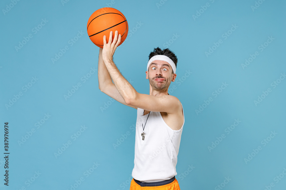 Loony man basketball player with thin skinny body sportsman in headband shirt whistle playing hold ball squinting eyes showing tongue isolated on blue background. Workout gym sport motivation concept.