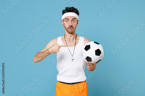 Perplexed fitness man football player with thin skinny body sportsman in headband shirt shorts whistle pointing finger on soccer ball isolated on blue background. Workout gym sport motivation concept. © ViDi Studio