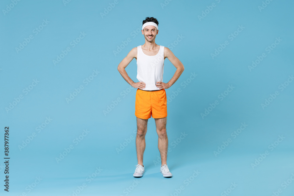Full length portrait smiling young fitness man with skinny body sportsman in headband shirt shorts standing with arms akimbo on waist isolated on blue background. Workout gym sport motivation concept. Full length portrait smiling young fitness man with skinny body sportsman in headband shirt shorts standing with arms akimbo on waist isolated on blue background. Workout gym sport motivation concept.
