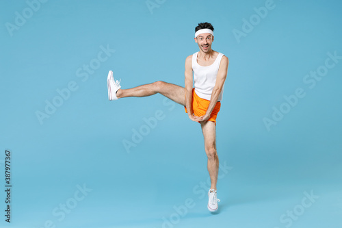 Full length portrait of cheerful young sporty fitness man with skinny body sportsman in white headband shirt shorts rising leg isolated on blue background studio. Workout gym sport motivation concept.