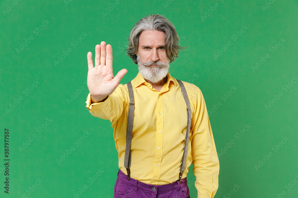 Displeased elderly gray-haired mustache bearded man wearing casual yellow shirt suspenders showing STOP gesture with palm looking camera isolated on bright green colour background, studio portrait.