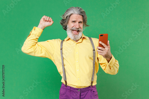 Joyful elderly gray-haired mustache bearded man in yellow shirt suspenders using mobile cell phone typing sms message doing winner gesture isolated on bright green colour background studio portrait. © ViDi Studio