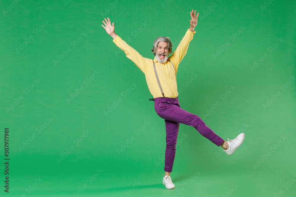 Full length of excited cheerful elderly gray-haired mustache bearded man in yellow shirt suspenders dancing rising spreading hands and legs isolated on bright green colour background studio portrait.