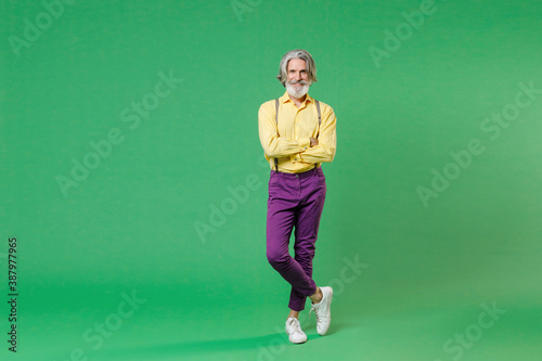 Full length of smiling elderly gray-haired mustache bearded man wearing casual yellow shirt suspenders holding hands crossed looking camera isolated on bright green colour background, studio portrait. © ViDi Studio
