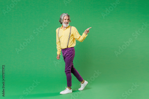 Full length side view of smiling elderly gray-haired mustache bearded man in casual yellow shirt suspenders pointing index finger aside isolated on bright green colour background, studio portrait. © ViDi Studio