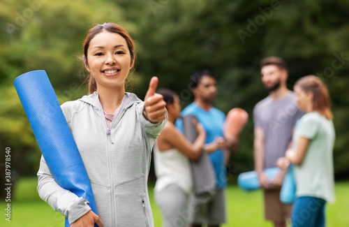fitness, sport and healthy lifestyle concept - happy smiling young asian woman with mat showing thumbs up over group of people meeting for yoga class at summer park