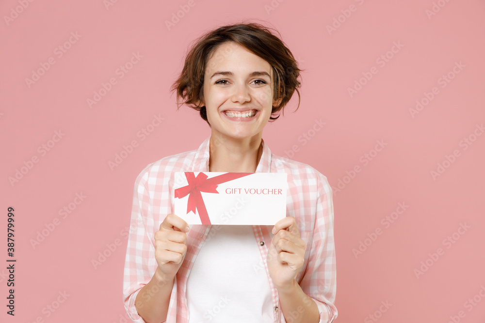 Smiling funny cheerful attractive beautiful young brunette woman 20s wearing casual checkered shirt standing hold in hands gift certificate isolated on pastel pink colour background, studio portrait. Smiling funny cheerful attractive beautiful young brunette woman 20s wearing casual checkered shirt standing hold in hands gift certificate isolated on pastel pink colour background, studio portrait.