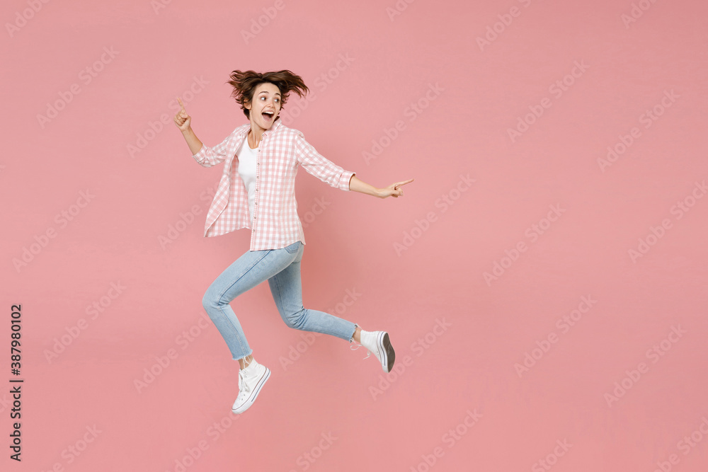 Full length side view of excited funny shocked young brunette woman 20s wearing casual checkered shirt jumping pointing index finger aside isolated on pastel pink colour background, studio portrait.