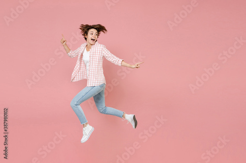 Full length side view of excited funny shocked young brunette woman 20s wearing casual checkered shirt jumping pointing index finger aside isolated on pastel pink colour background, studio portrait.