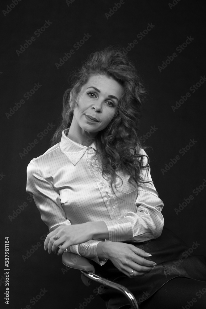 A beautiful slender brunette with long curly hair in a white satin blouse sits in the studio on a black background, bw