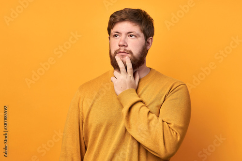Image of a man in basic clothing, thinking and touching his chin, looking to the side, isolated on a yellow background. The bearded man makes a decision. to be or not to be