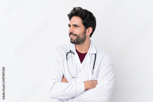 Young handsome man with beard over isolated white background wearing a doctor gown and with arms crossed looking side