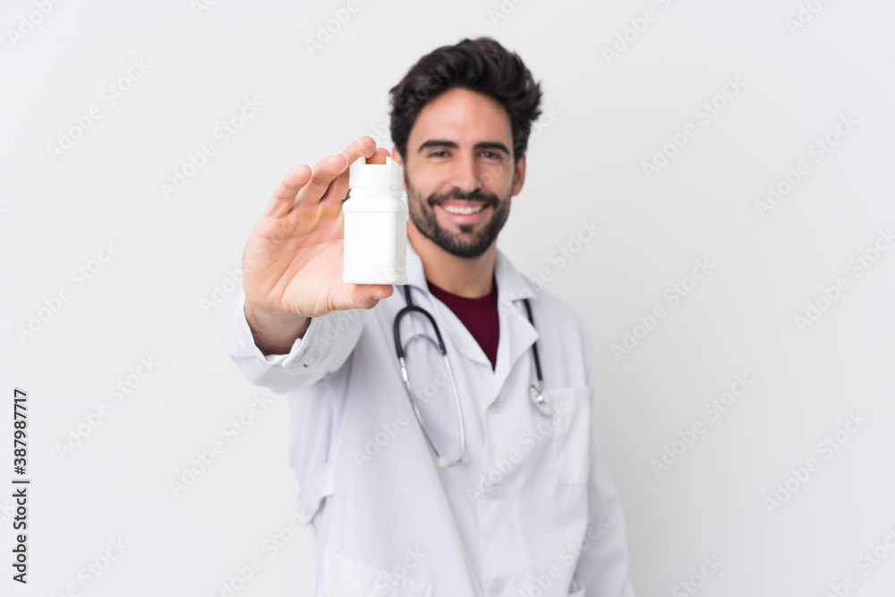 Young handsome man with beard over isolated white background wearing a doctor gown and holding pills