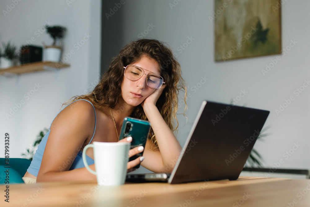 Bored young woman working at home with laptop and using smartphone. Bored young woman working at home with laptop and using smartphone.