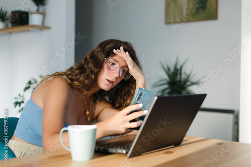 Expressive incredulous young woman using laptop and smartphone at home. Working at home issues concept.