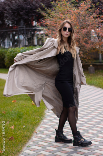 attractive slim young brunette in fashionable trench, black dress and sunglasses in park. autumn, yellow leaves. © OliaVesna