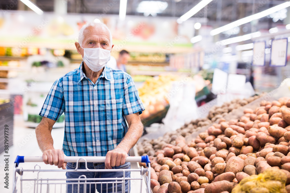 mature caucasian man in mask with covid protection choosing potatoes in vegetable section of supermarke