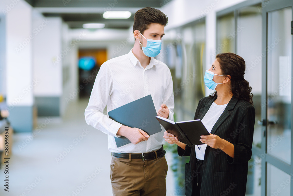 Two colleagues in protective face masks in modern office discussing together work issues. Business People back at work in office after quarantine. Covid-19.