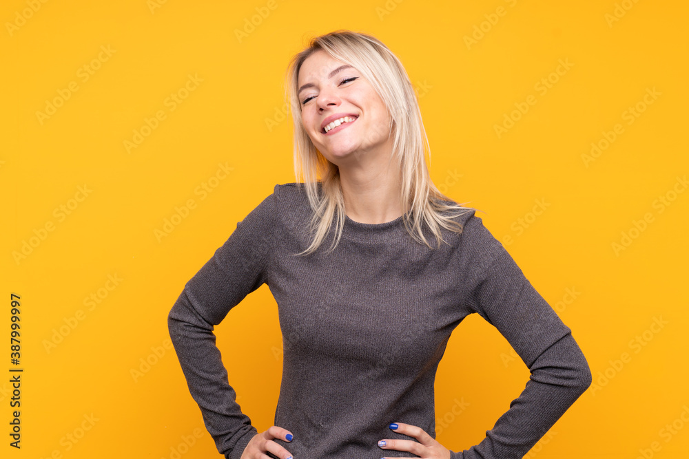 Young blonde woman over isolated yellow background posing with arms at hip and smiling