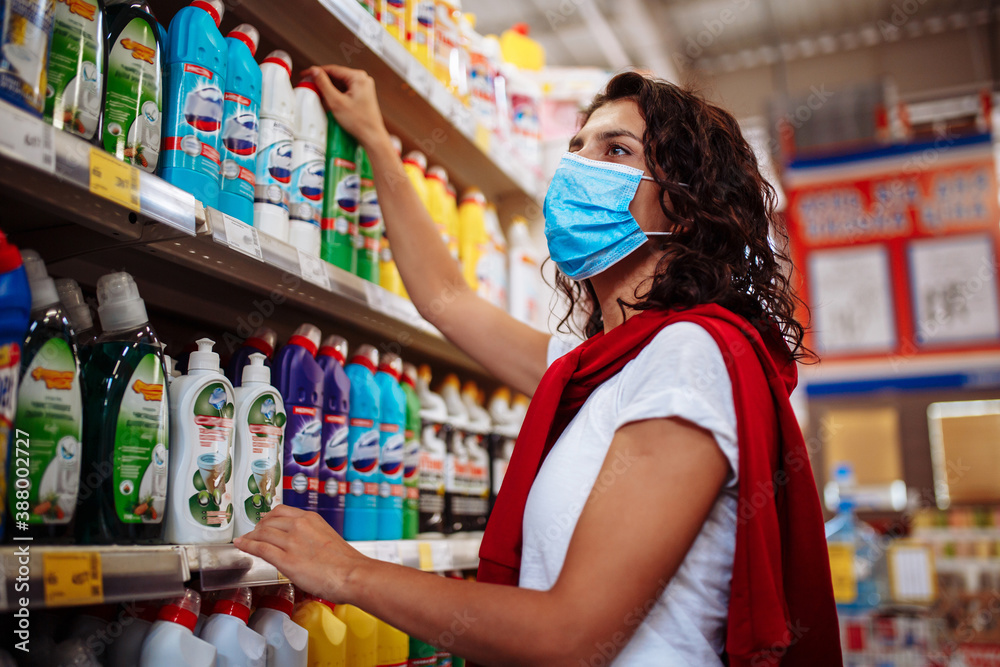 Young woman picks up the disinfecting and cleaning scour at a supermarket wearing a medical sterile mask during the coronavirus pandemic quarantine. Healthcare and home sanitizing concept.