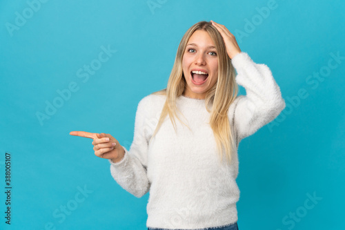 Young blonde woman isolated on blue background surprised and pointing finger to the side