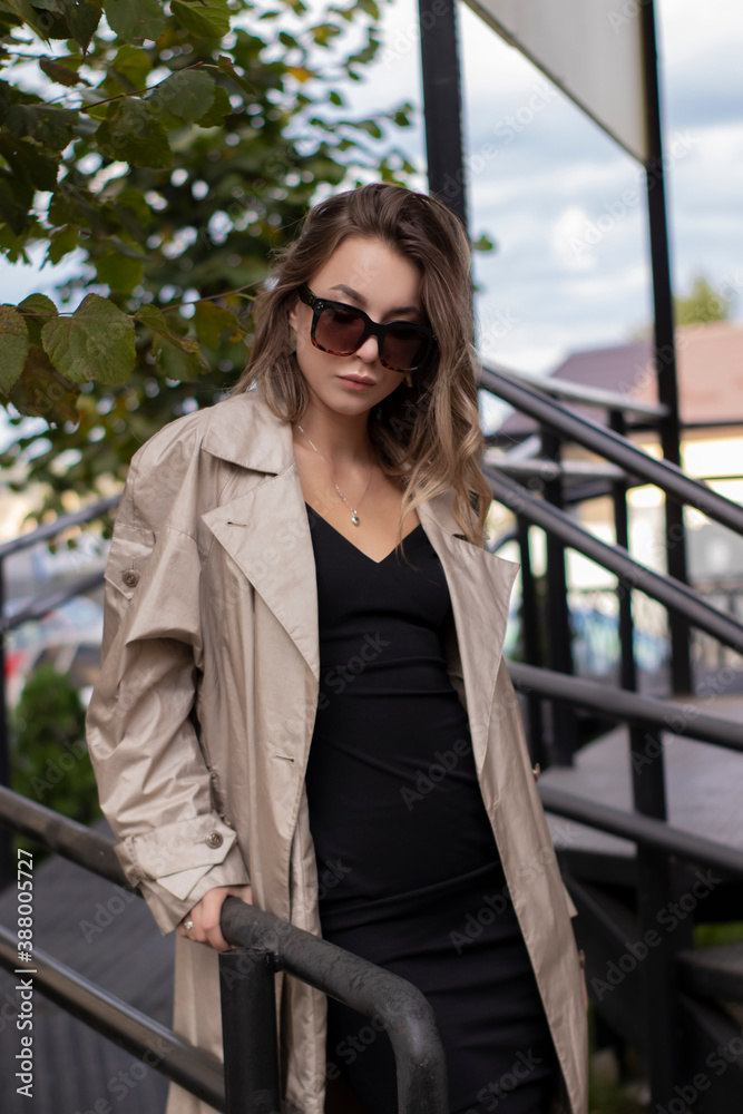 beautiful slim young brunette in fashionable trench coat, black dress and sunglasses walking near stairs. autumn, yellow leaves.