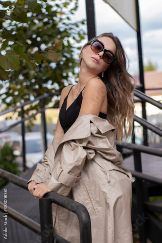 beautiful slim young brunette in fashionable trench coat, black dress and sunglasses walking near stairs. autumn, yellow leaves.