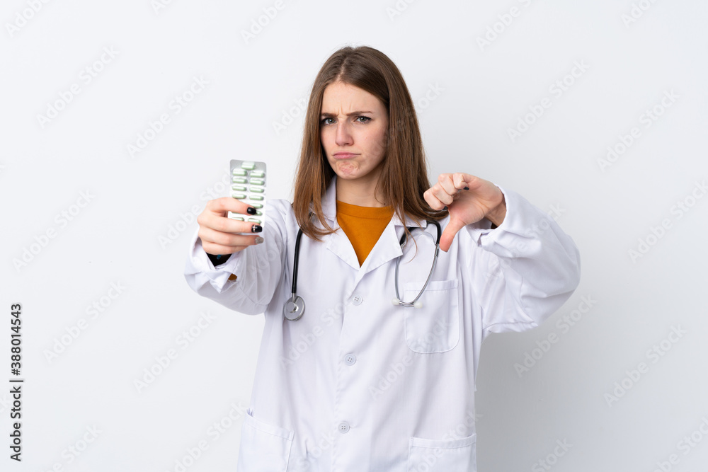 Young woman over isolated background wearing a doctor gown and holding pills while doing bad signal Young woman over isolated background wearing a doctor gown and holding pills while doing bad signal