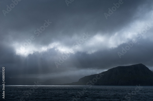 Sailing by Suðuroy island in dramatic light. Moody clouds on sky and sun rays (god rays) coming through and lighting up the ocean. Faroe Islands.